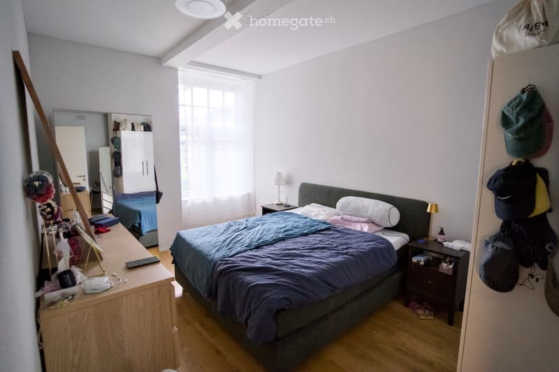 Cozy bedroom with a double bed, nightstands, a mirror, and a wardrobe. The room features wooden flooring and large windows with blinds.
