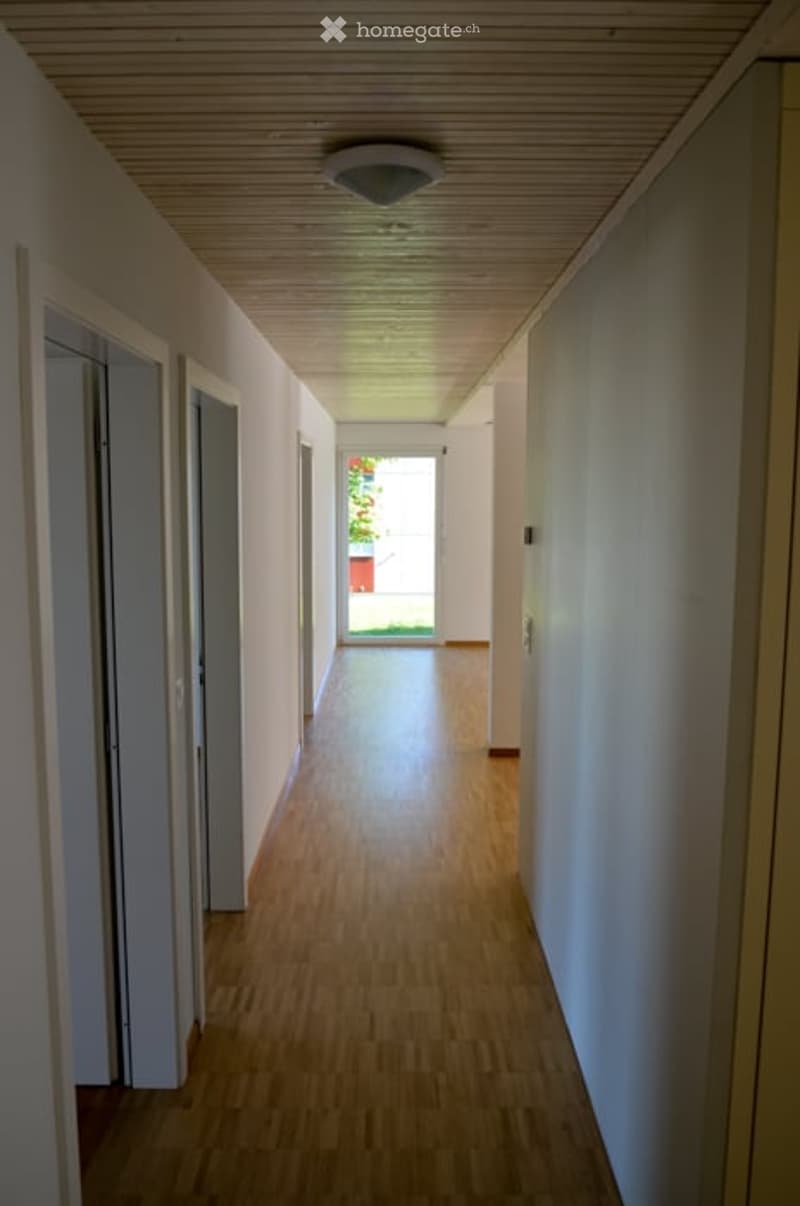 A long, narrow hallway with light wooden flooring and white walls, featuring a recessed ceiling light and several doors on both sides. Natural light streams in from an open doorway at the end, revealing a glimpse of greenery outside.