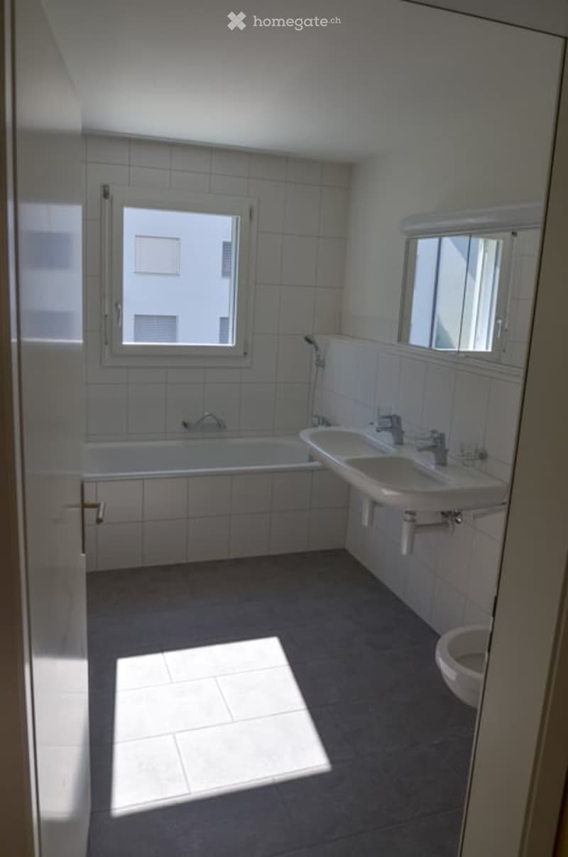 Bright bathroom with white tiled walls and floor, featuring a bathtub, sink, toilet, and a large mirror above the sink. Natural light streams in from a window.