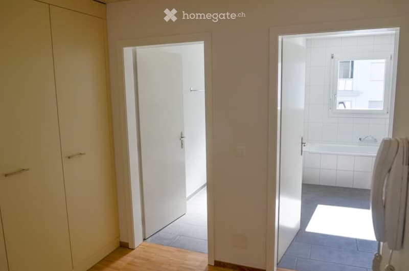 Empty room with wooden flooring, white walls, and open doors leading to a bathroom and another room.