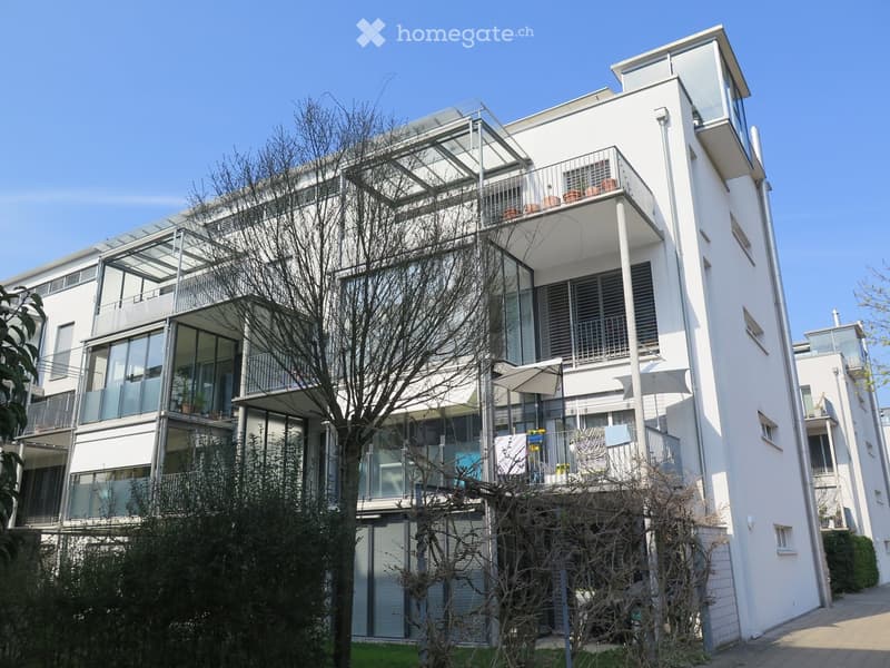 Modern multi-story residential building with white facade, large windows, balconies, and outdoor seating areas.