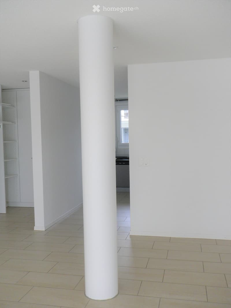 Empty room with tiled floor, white walls, and a large white column in the center. A doorway leads to another room with a visible kitchen area.