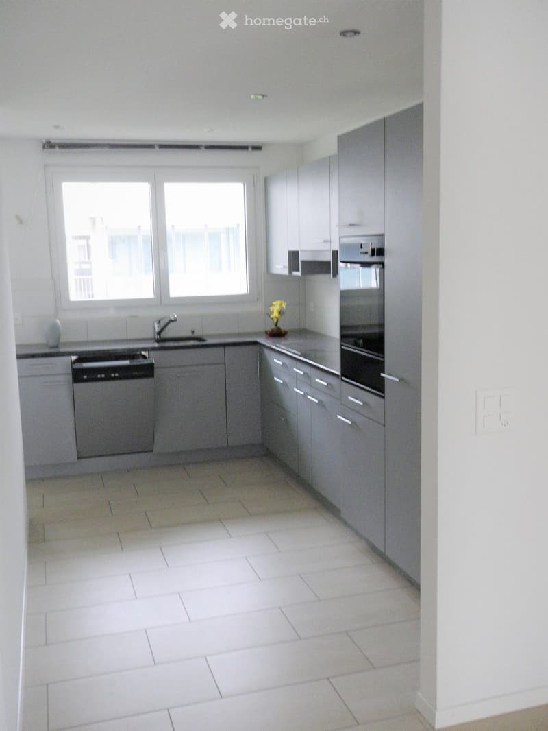 Modern kitchen with white tiled floor, grey cabinets, sink, dishwasher, oven, and a small flower vase on the counter.