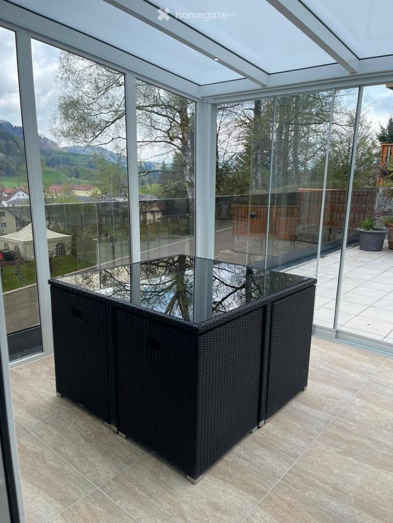Glass-enclosed balcony with a wicker sofa, large windows offering a scenic view of the landscape.