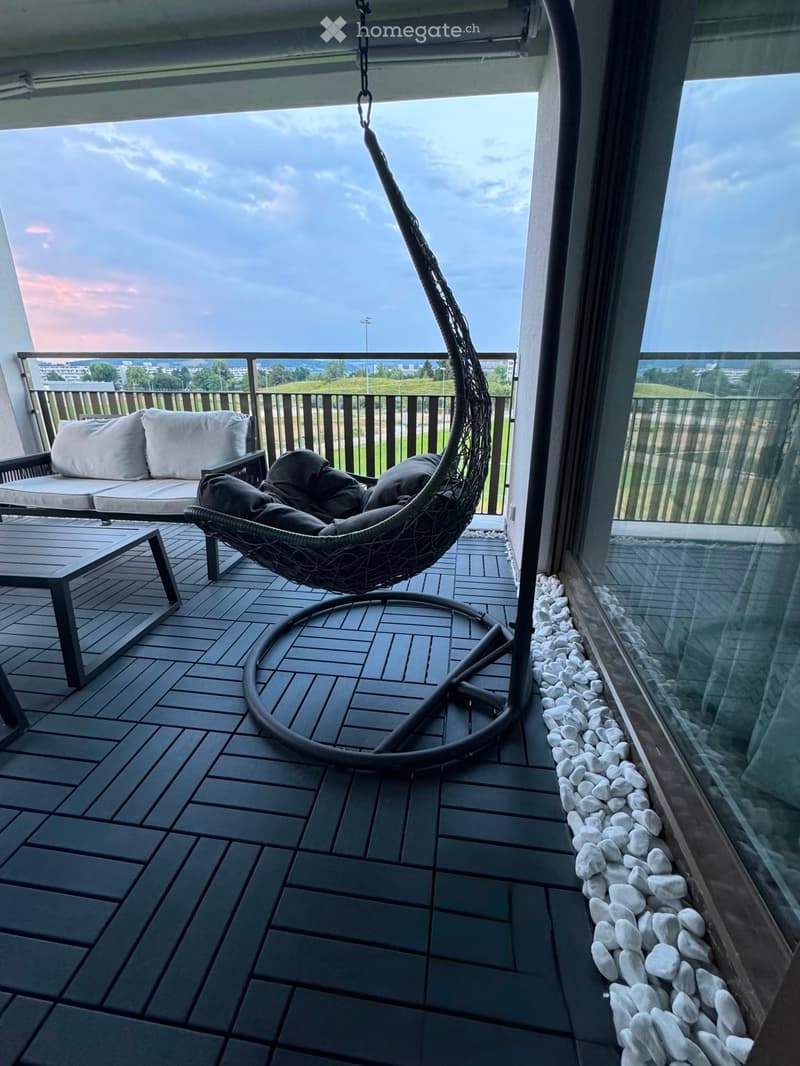 Balcony with a wicker hanging chair, a small sofa, and a table, overlooking a scenic view.
