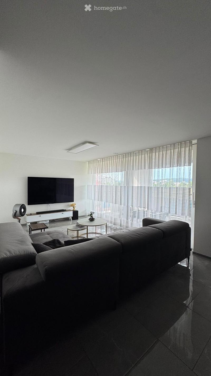 Modern living room with a grey sectional sofa, coffee table, television, fan, and small round table near large windows with sheer curtains.