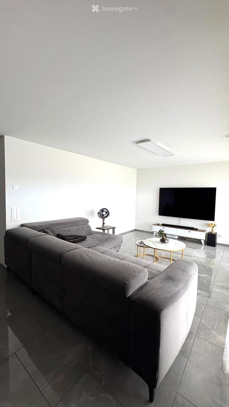 Modern living room with a grey sectional sofa, a white coffee table, a flat-screen TV mounted on the wall, and a small side table with a fan.