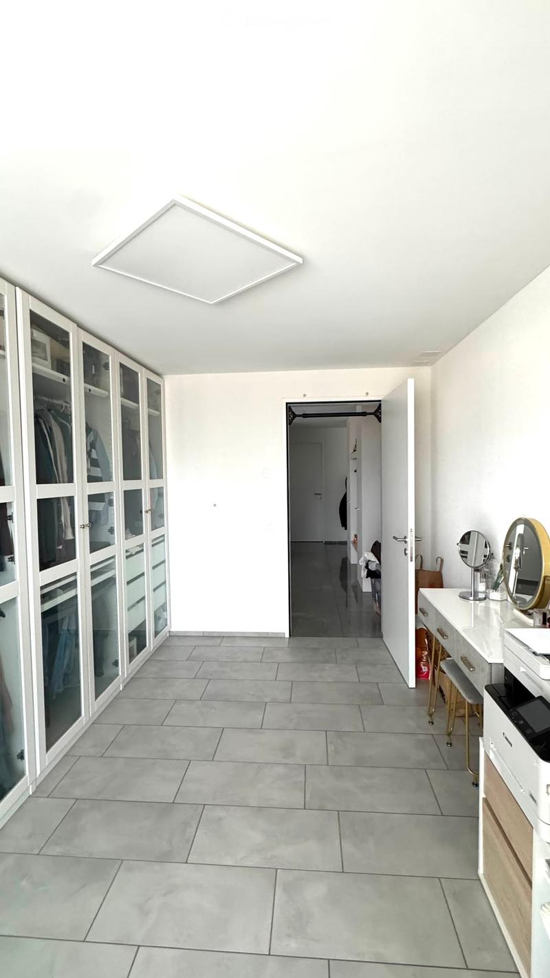 Bright, minimalist hallway with grey tiled flooring, white walls, and a large recessed ceiling light. Features built-in mirrored wardrobes on one side and a vanity area with a mirror, stool, and printer on the other.