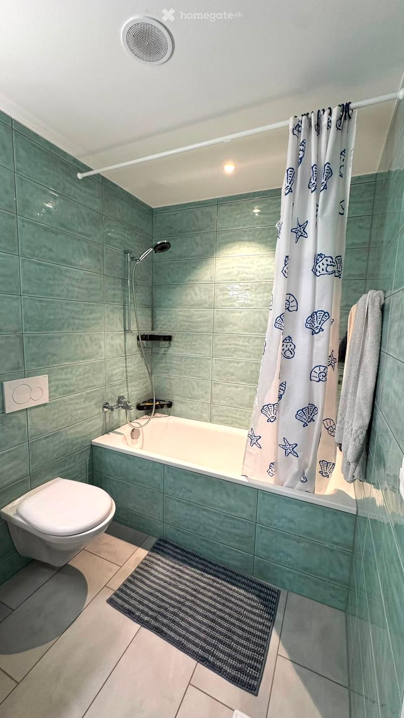 Modern bathroom with green tiled walls, a bathtub, toilet, shower curtain with sea-themed design, and a towel hanging on the side.