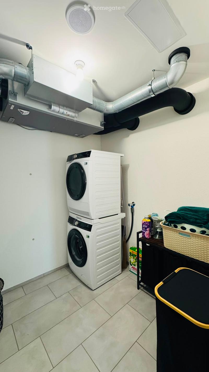 A small utility room with a stacked washing machine and dryer, a small table with a basket, and various cleaning supplies.