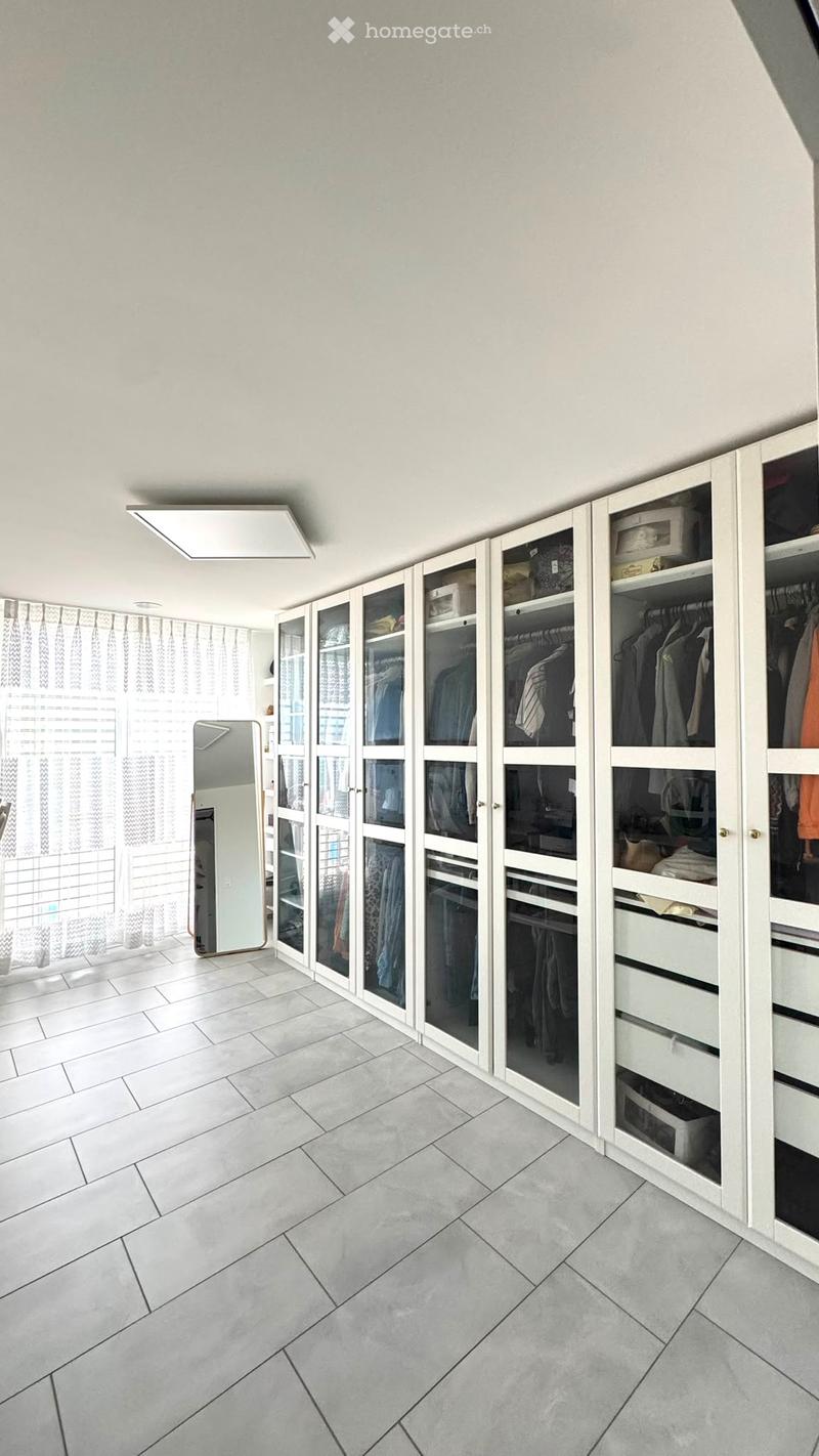 A spacious, well-lit walk-in closet with white built-in wardrobes, glass-fronted sections, and a large mirror on the wall.
