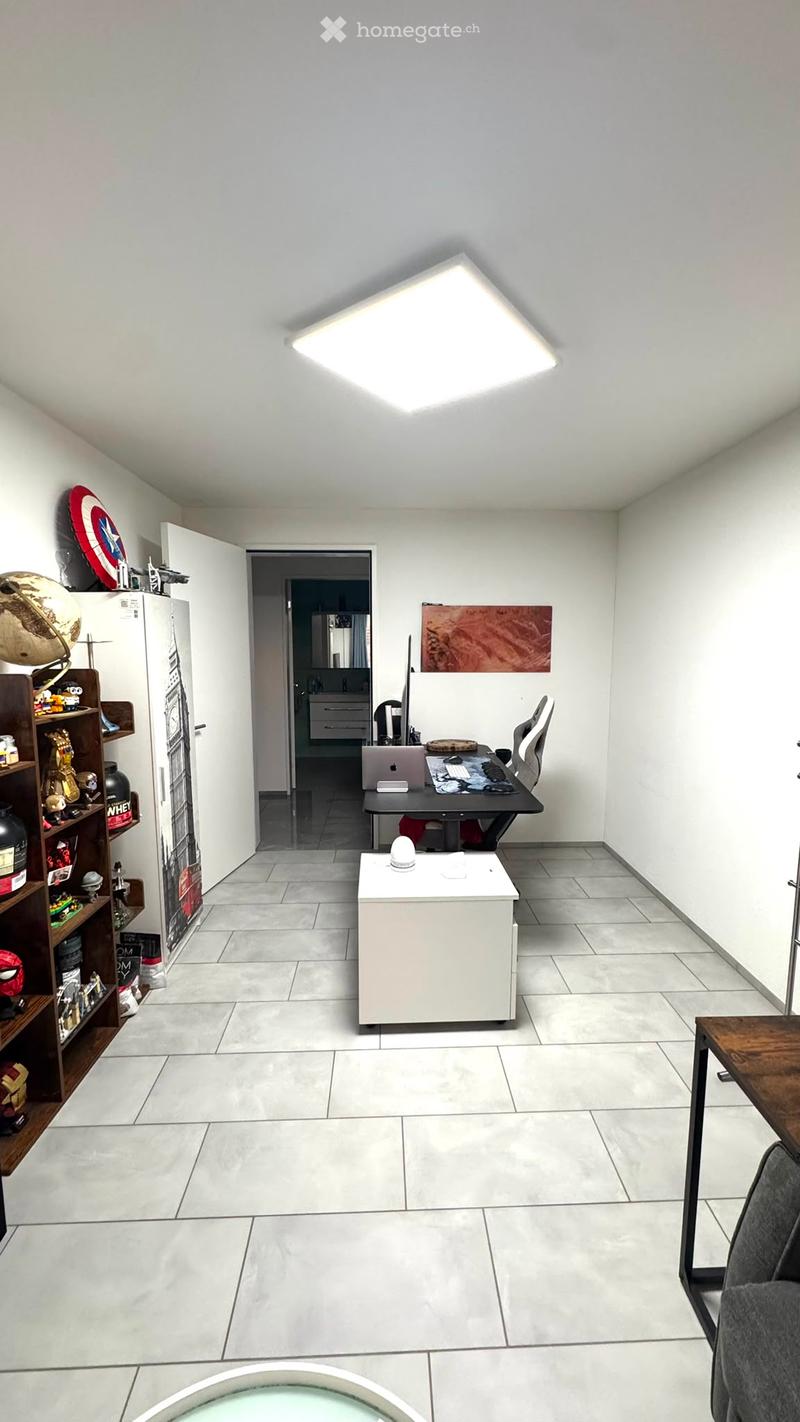 A modern, well-lit room with a desk setup, a small table, and a shelf with various items. The room features white walls, grey tiles, and a large square ceiling light.