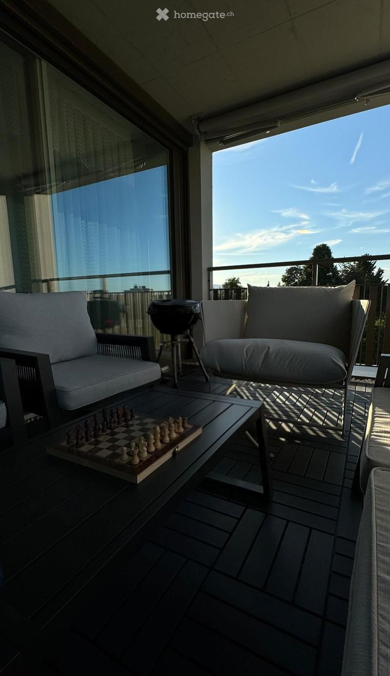Modern balcony with dark tiled floor, white sofa, coffee table, chessboard, and grill, overlooking a cityscape with clear sky.
