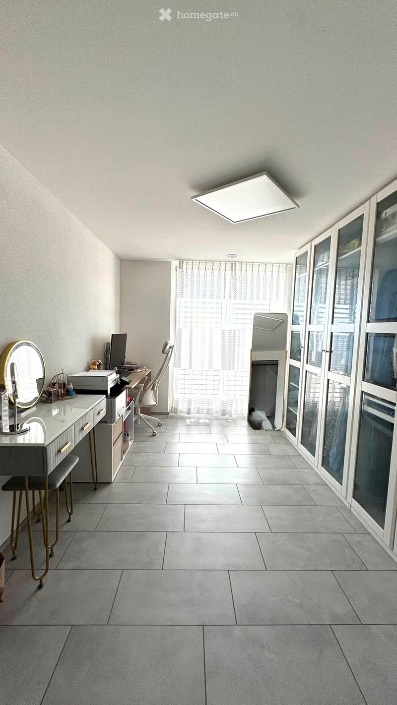 A modern, well-lit room with a vanity area, a desk, and a chair. The space features large windows with sheer curtains and a sleek, minimalist design.