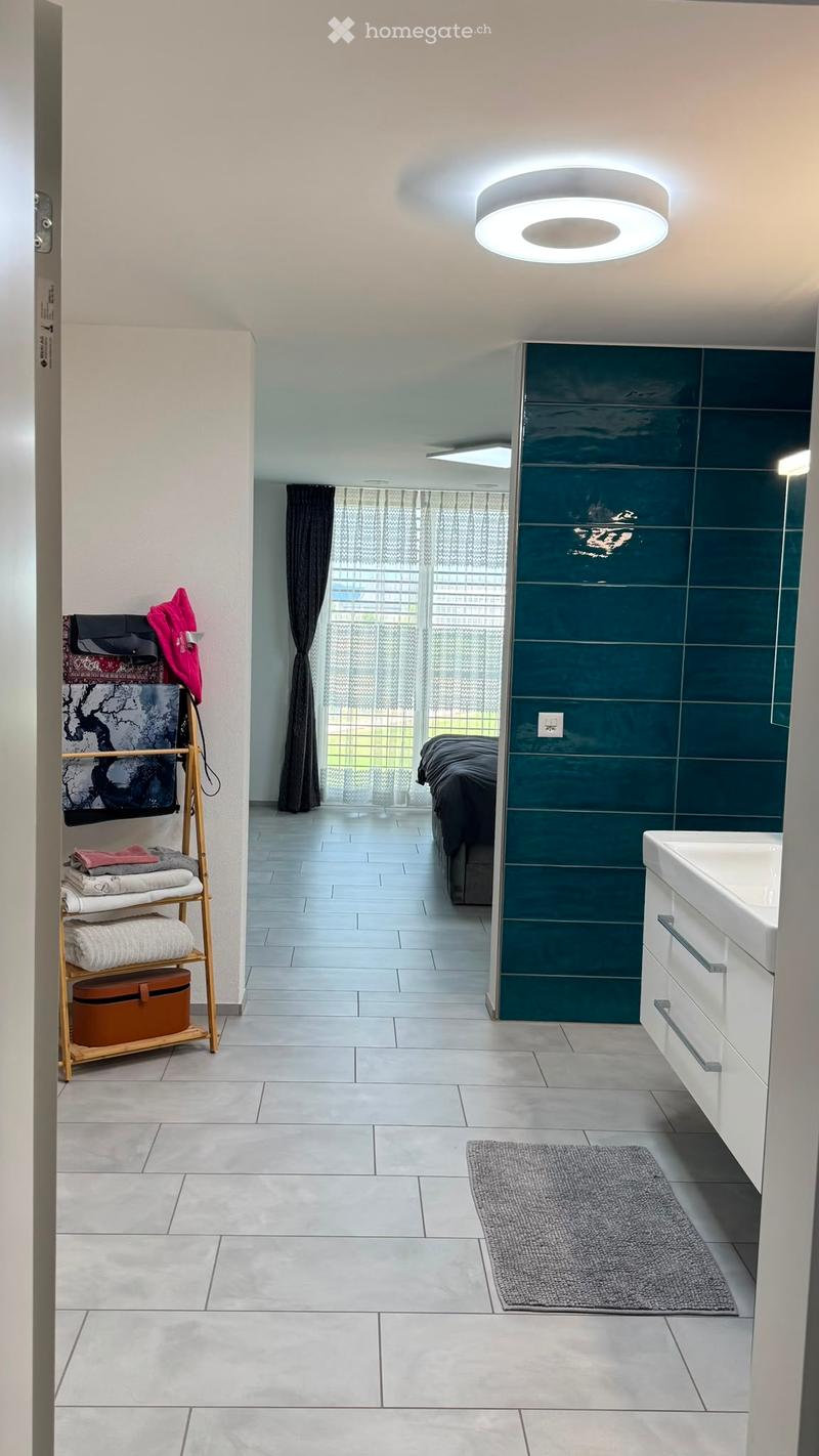 Modern bathroom with white tiles, a large sink, a mirror, a towel rack with folded towels, a bed, and a grey rug.