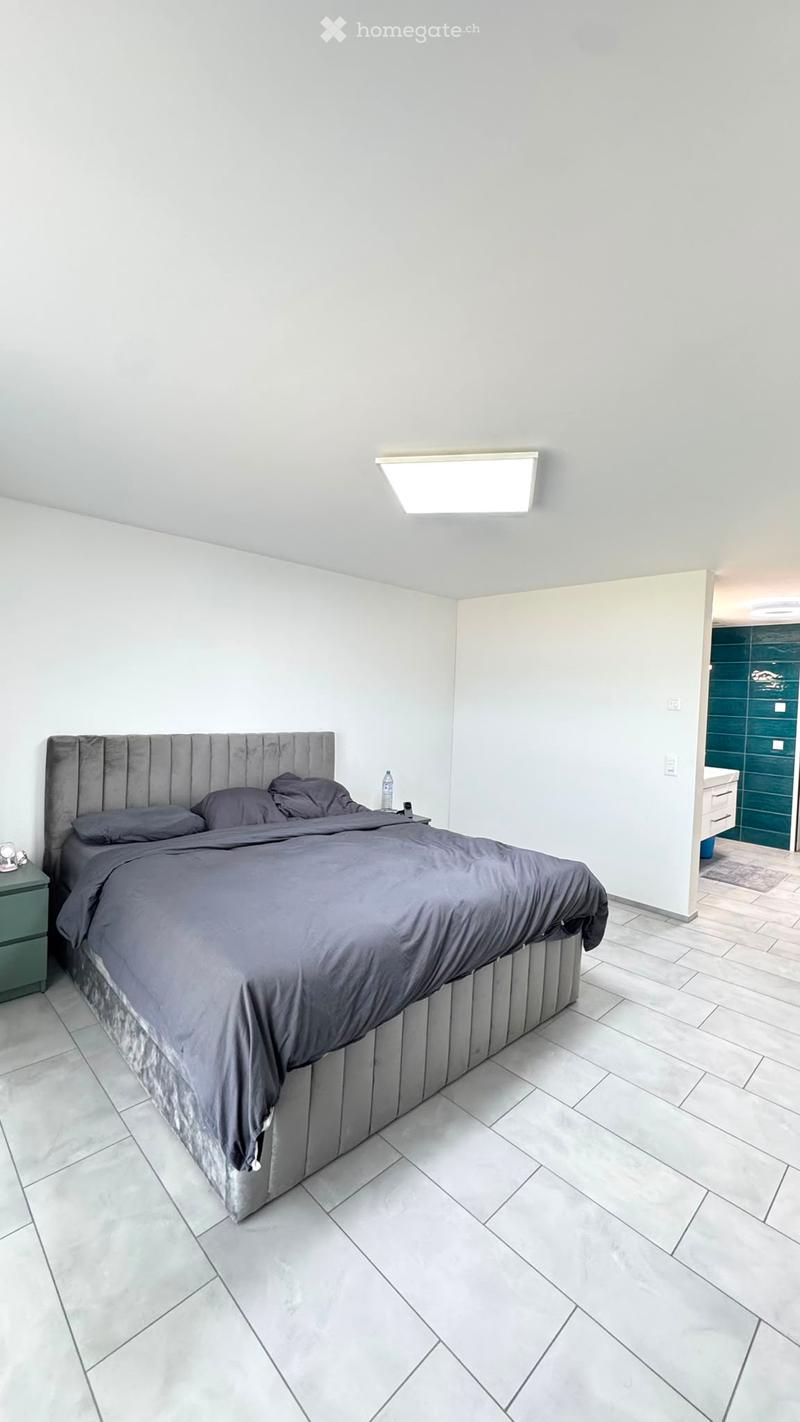 Modern bedroom with a large bed, nightstand, and a bottle on the nightstand. The room features light grey walls, a tiled floor, and a built-in wardrobe.