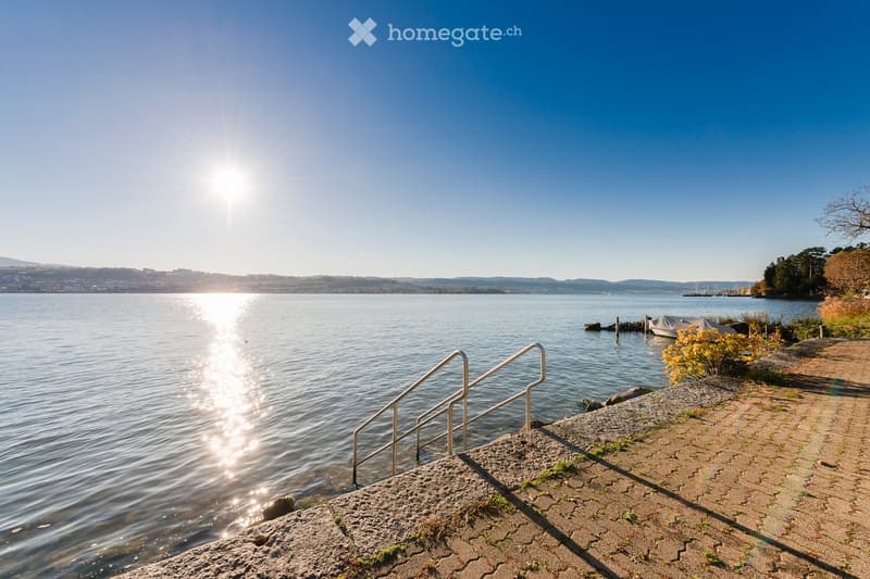 A serene lakeside scene with a paved walkway leading down to steps that descend into the water, surrounded by calm lake waters and distant hills under a clear blue sky.
