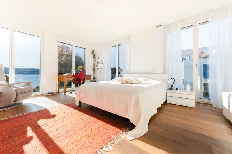Bright bedroom with large windows offering a scenic view, featuring a bed with white bedding, a red accent chair, a wooden desk with a chair, and a nightstand with a lamp.