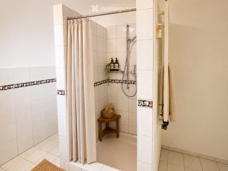 Modern bathroom with a shower area featuring a curtain, a small wooden stool with a basket, and a towel rack with a towel. The shower has a glass door and a decorative tile border.