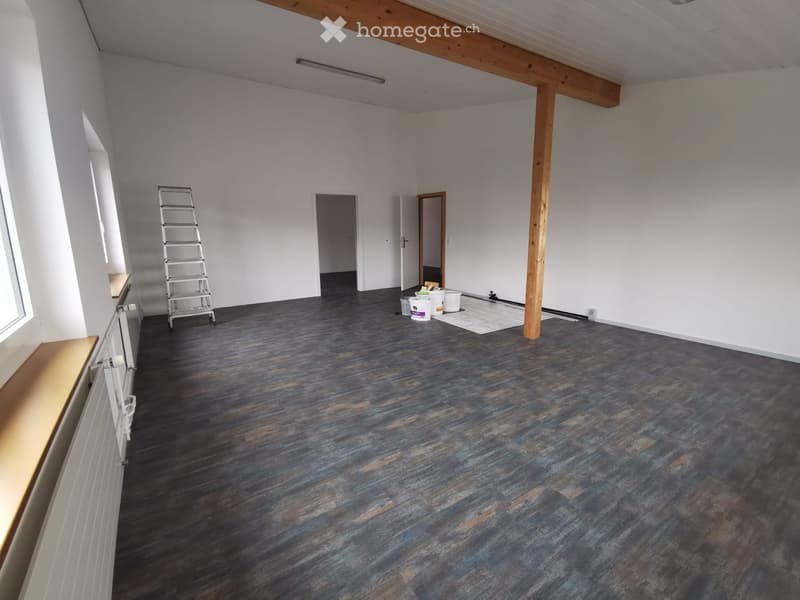 Empty room with white walls, dark wooden flooring, and exposed wooden beams on the ceiling. A ladder and some construction materials are present, indicating ongoing work.