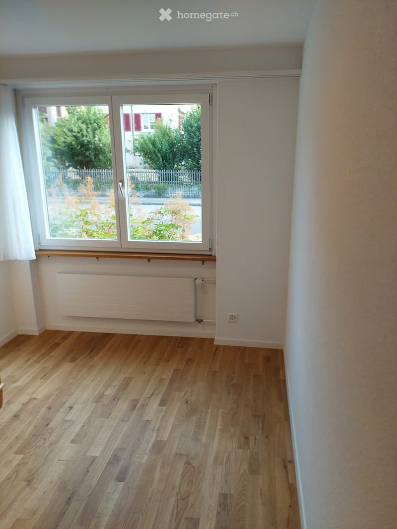 Empty room with wooden flooring, white walls, large window with white frame, radiator, and white curtain.