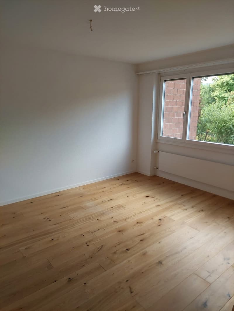 Empty room with light wooden flooring and white walls, featuring a large window with a view of greenery outside.