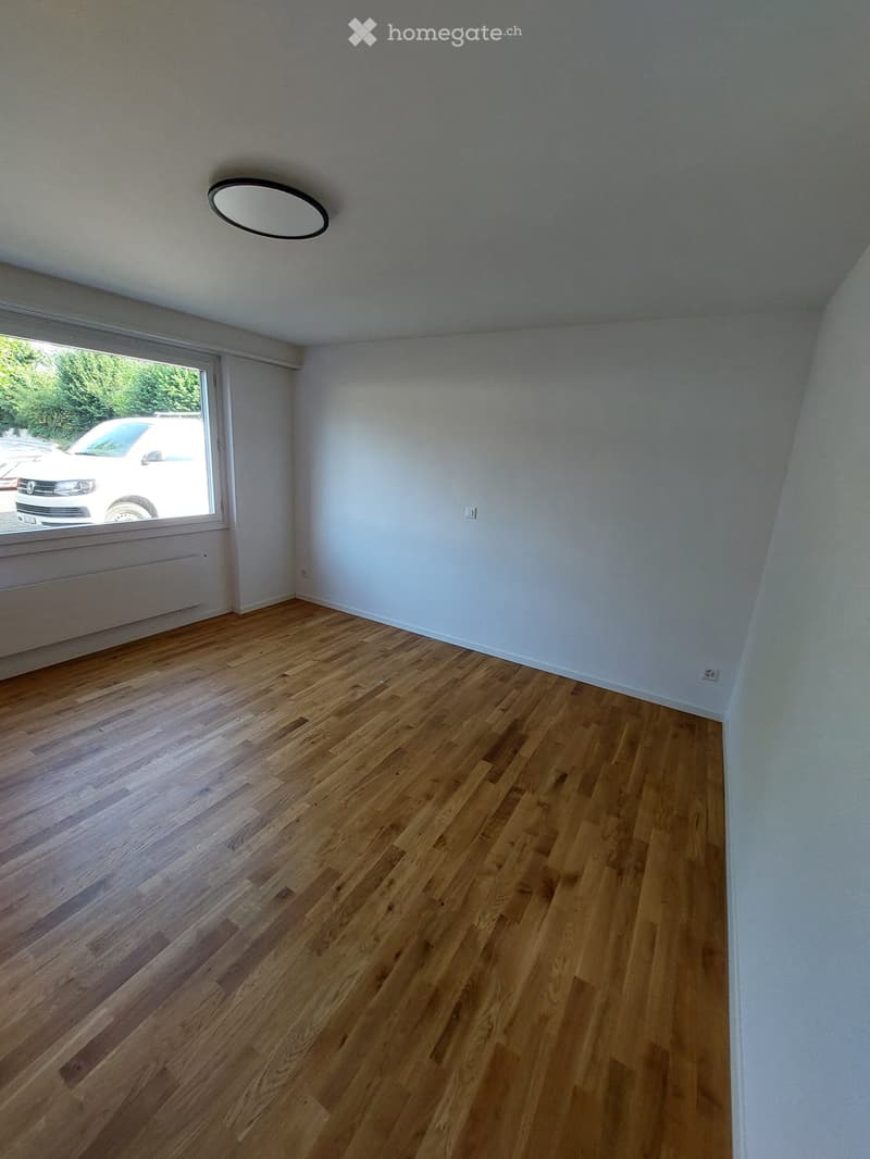 Empty room with wooden flooring, white walls, and a large window with a view of a parked car outside.