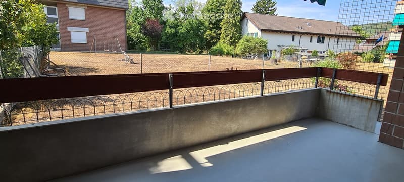 Empty balcony with concrete floor, wooden railing, and metal fence, overlooking a fenced yard with trees and neighboring houses.