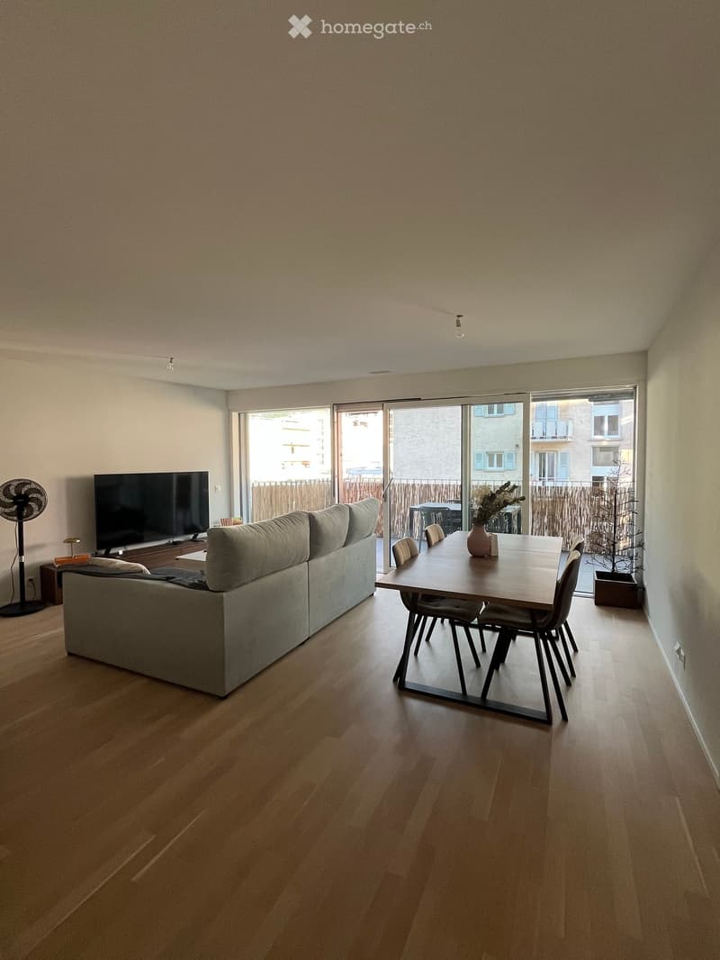 Open-plan living and dining area with wooden flooring, grey sectional sofa, dining table with chairs, TV, and fan.
