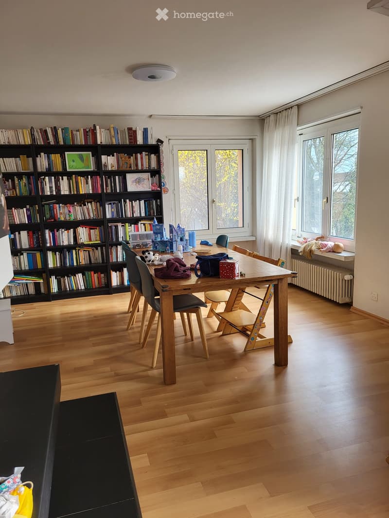 Bright room with wooden flooring, large windows, bookshelf filled with books, dining table with chairs, and toys scattered around.