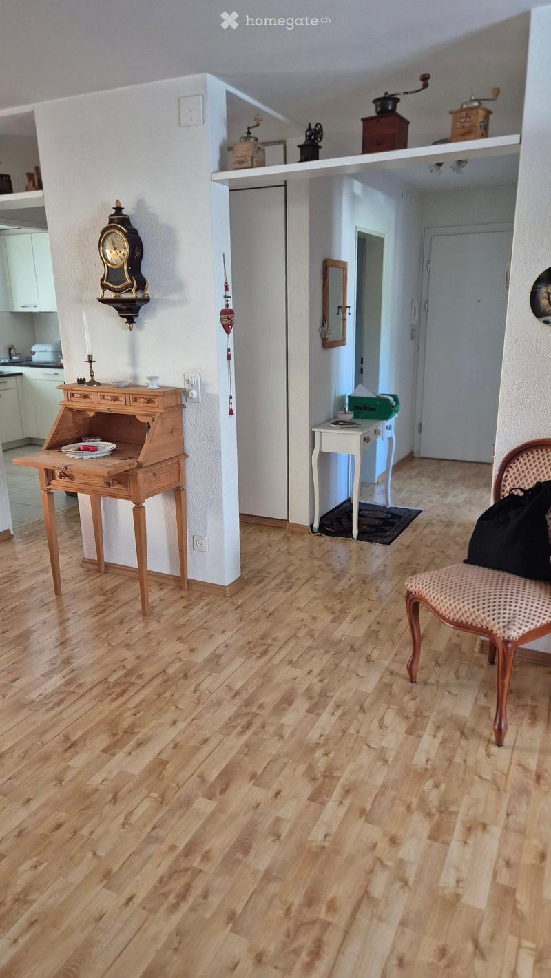 Hallway with wooden flooring, white walls, and several doors leading to different rooms. Visible items include a small wooden desk, a wall-mounted clock, a chair, and a small table with a mirror.