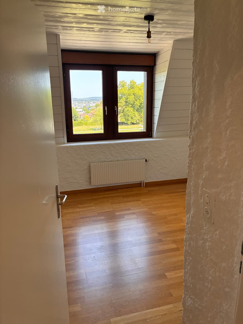 Empty room with wooden flooring, white walls, a radiator, and a large window with a view of greenery outside.