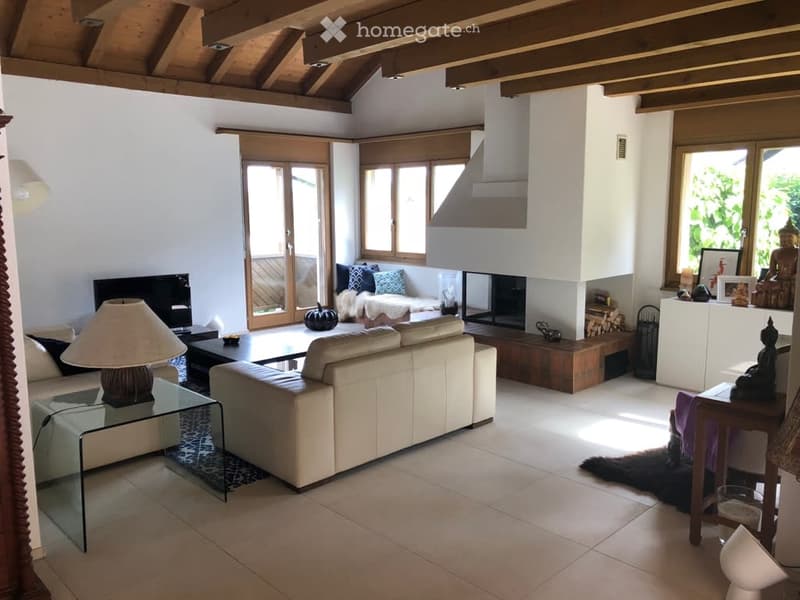 Modern living room with a fireplace, large windows, and a staircase leading to an upper level. Features include a cream-colored sofa, a glass coffee table, a floor lamp, and decorative items on a side table.