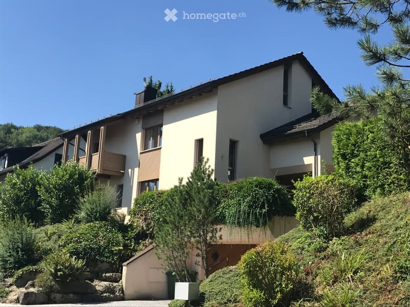 Two-story house with beige walls, dark roof, multiple windows, and a balcony on the upper level, surrounded by greenery and trees.