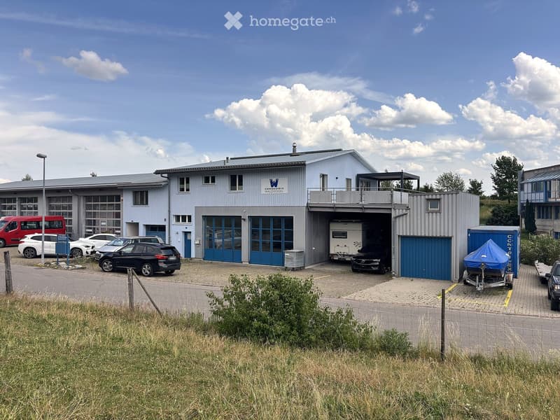 A modern, single-story building with a flat roof and large garage doors, surrounded by a paved parking area with several cars and a trailer.
