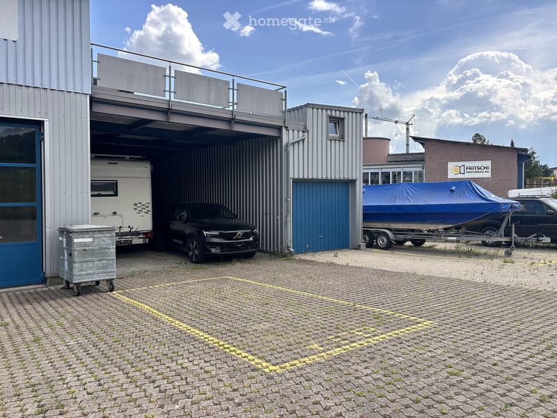 A modern industrial-style building with a large garage door open, showing a black car, a camper van, and a covered boat on a trailer inside. The building has a metal exterior and a raised balcony area.