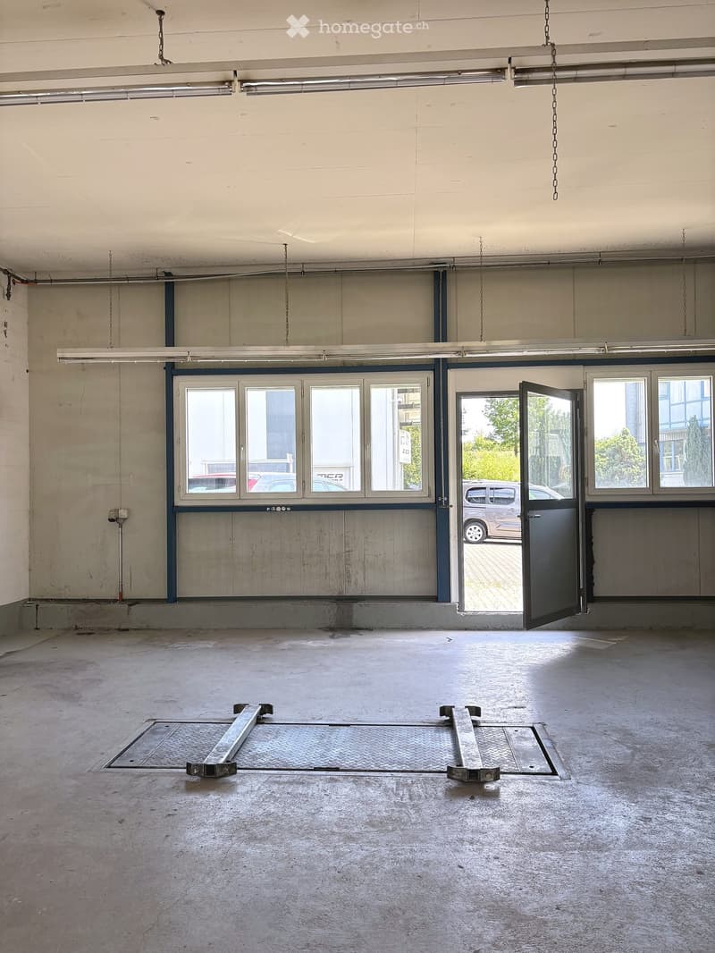 Empty garage with concrete floor, large windows, and a partially open door leading outside. No furniture or equipment present.