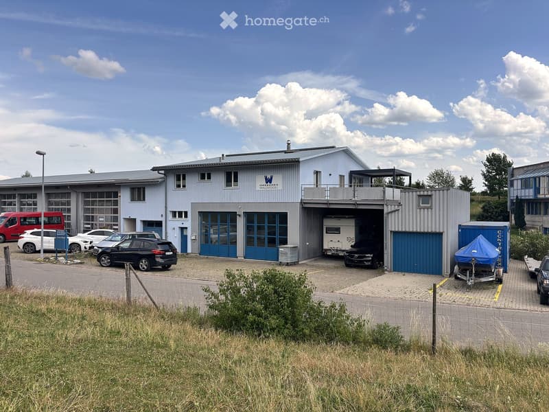 Modern industrial-style building with large garage doors, multiple parked cars, and a trailer.