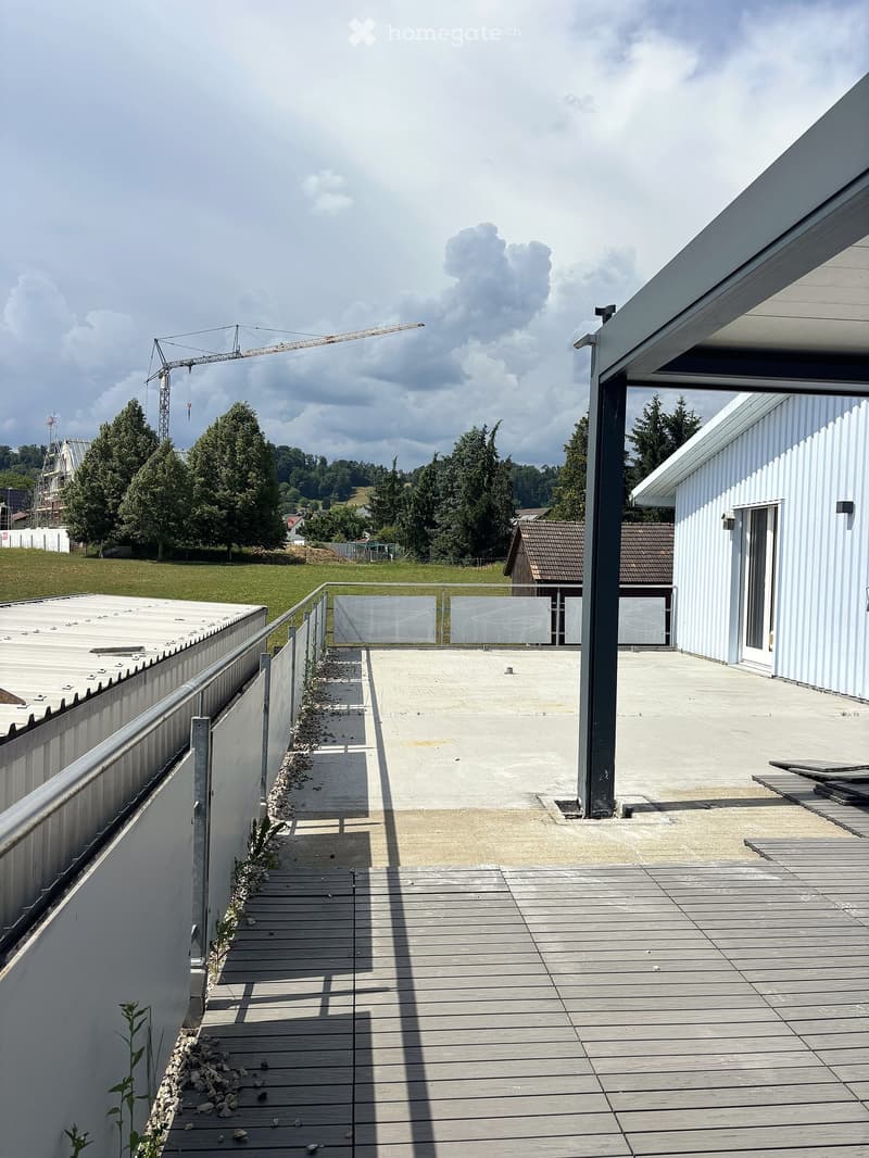Roof terrace with wooden decking, metal railing, and a view of a grassy area with trees and a construction crane in the background.