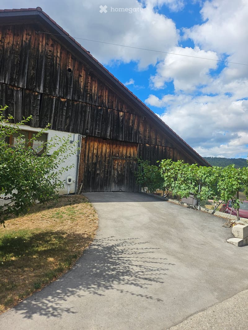 A large wooden barn with a red-tiled roof, surrounded by greenery and a gravel driveway.