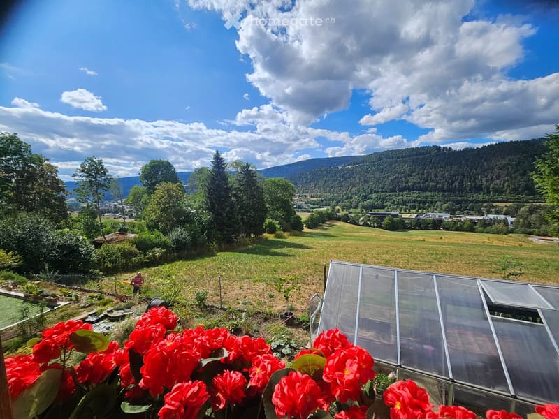 A scenic garden view with a greenhouse, red flowers in the foreground, and a panoramic view of a valley with hills and scattered houses.