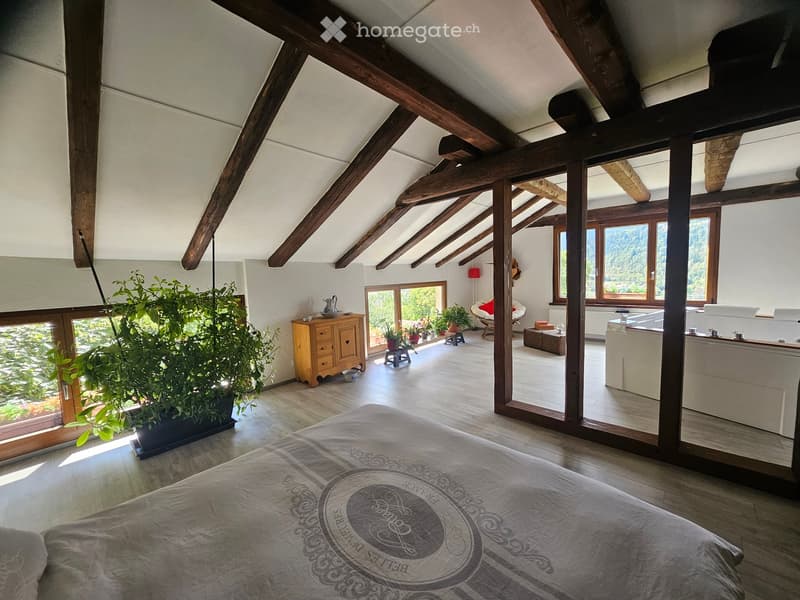 Bright, spacious bedroom with wooden beams, large windows offering a scenic view, a bed with patterned bedding, a wooden cabinet, a small table, a chair, and a bathtub in the adjacent area.