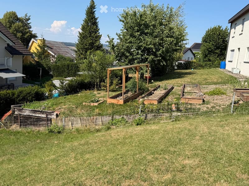 A well-maintained garden with raised wooden beds, a wooden pergola, and a fenced area. The garden is surrounded by trees and neighboring houses.