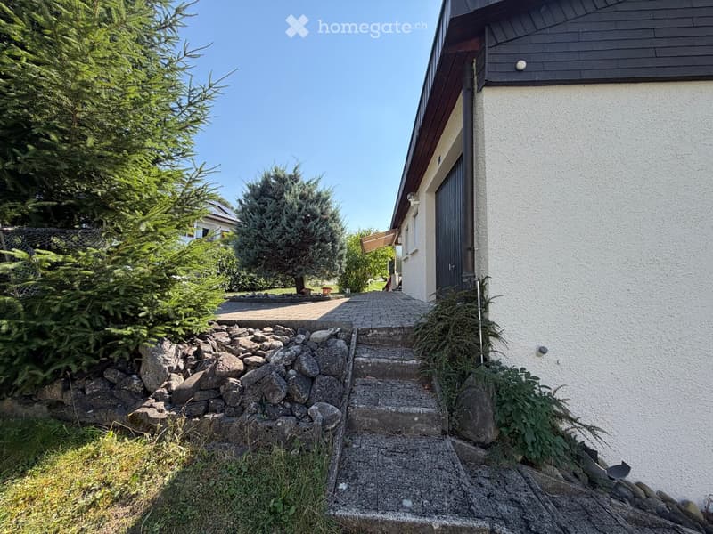 Garden area with stone steps leading to a house entrance, surrounded by greenery and rocks.