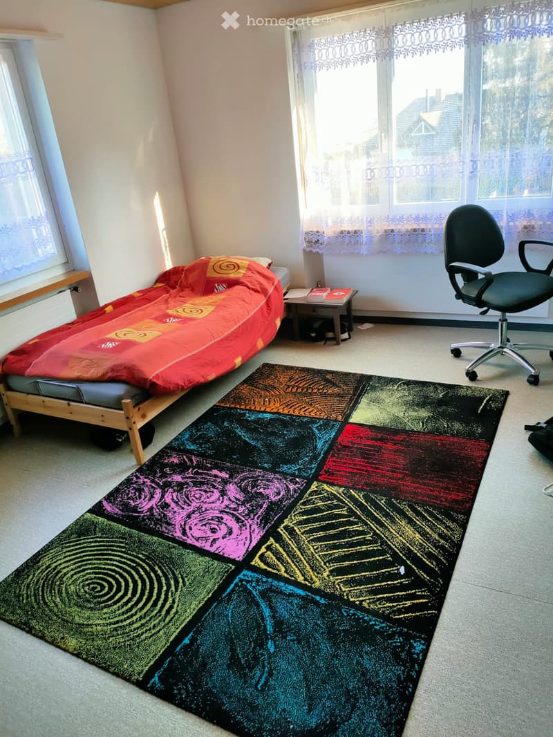 A small, cozy bedroom with a wooden bed, colorful bedspread, a colorful rug, a black office chair, a small table with books, and large windows with lace curtains.