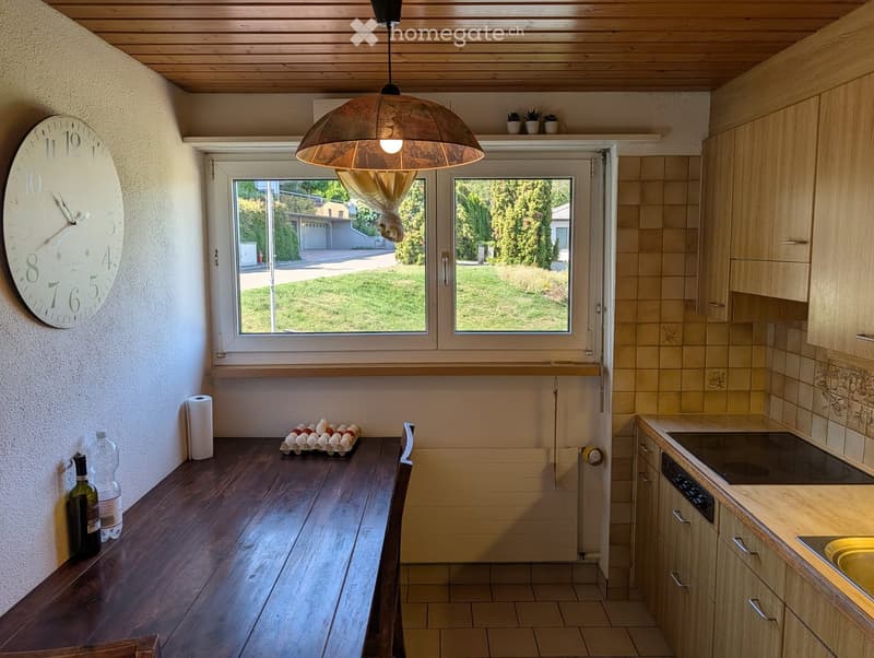 Bright kitchen with wooden cabinets, tiled backsplash, and a large window overlooking a green lawn. Features a wooden table with a bottle and eggs, a hanging light fixture, and a wall-mounted clock.