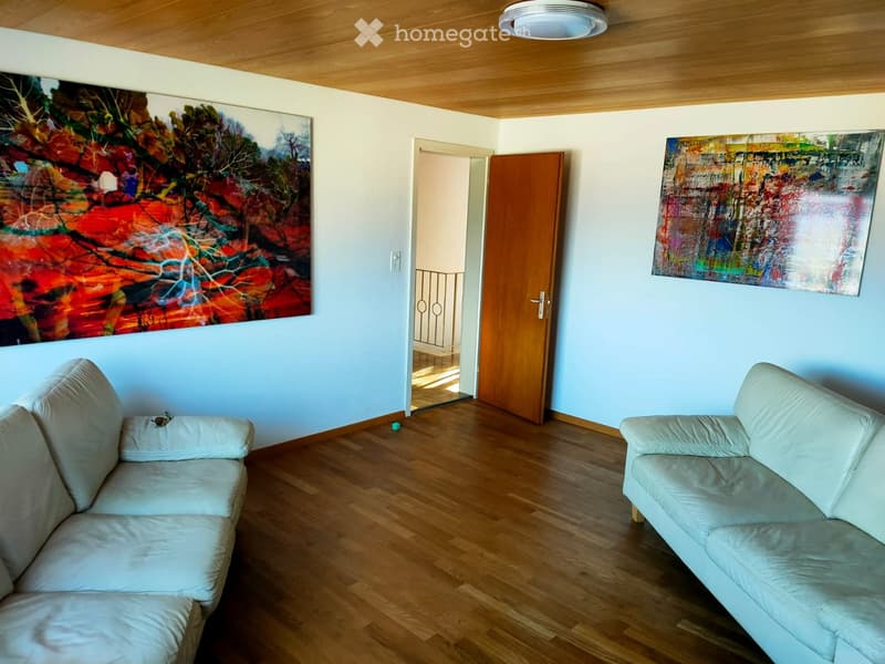 Living room with light-colored sofas, wooden flooring, and two large abstract paintings on the walls. A wooden door leads to another room.