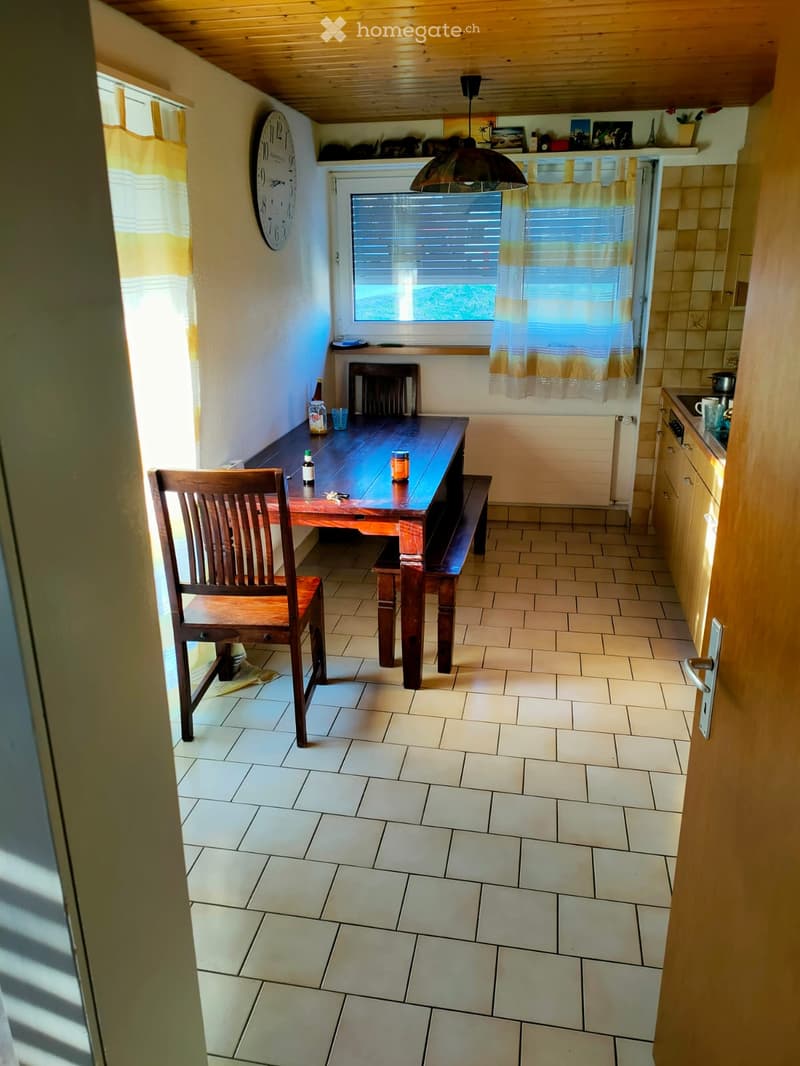 A small dining area with a wooden table, bench seating, and a single chair, located next to a kitchen area with cabinets and a window.