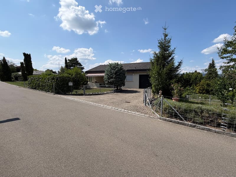 Single-story house with a dark garage door, surrounded by trees and shrubs, with a paved driveway and a metal fence.