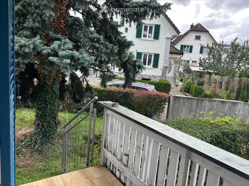 Balcony with wooden decking, white railing, metal handrail, and view of a residential area with houses, trees, and parked car.